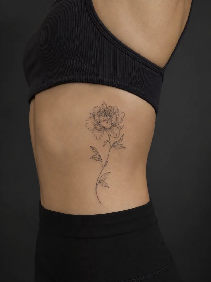 Fine-line peony tattoo - An open peony and curved stem follow the side torso.