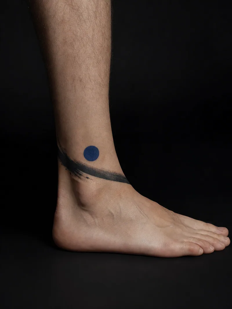Abstract ankle tattoo - sweeping black brushstroke paired with one cobalt-blue circle