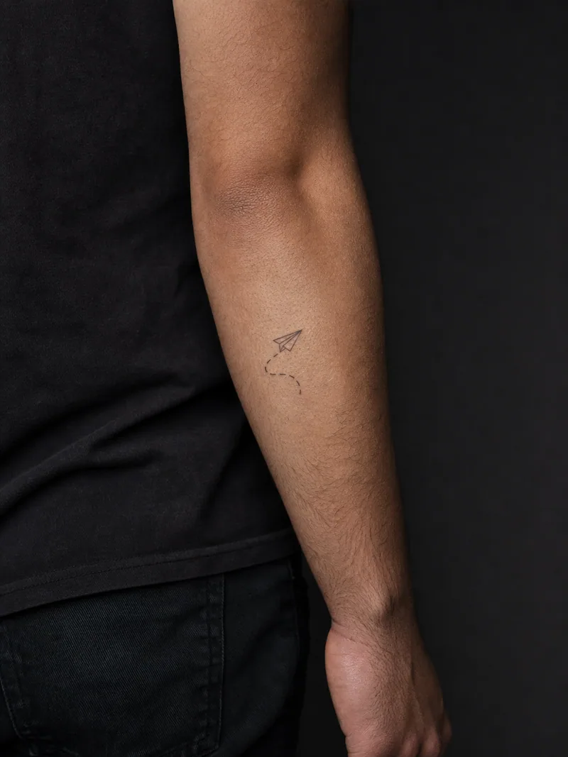 Paper airplane tattoo - A tiny paper plane follows a short broken flight path across the forearm.