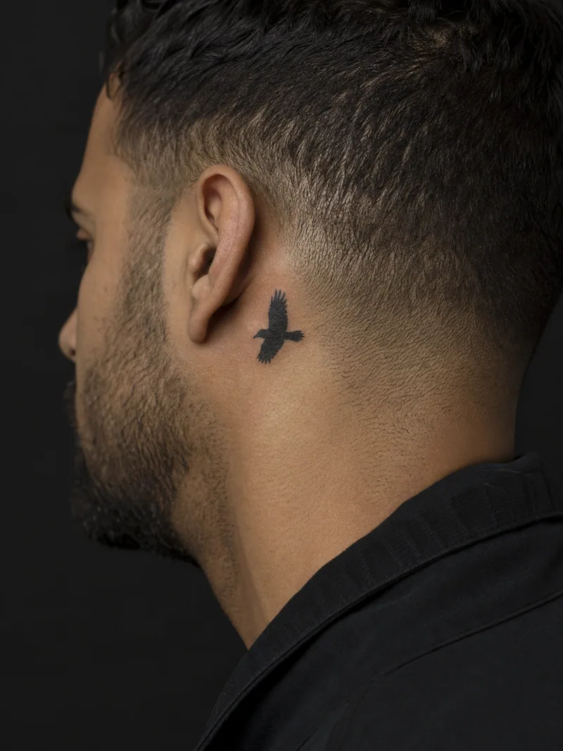 Raven silhouette tattoo - A flying raven reduces to a crisp winged silhouette behind the ear.
