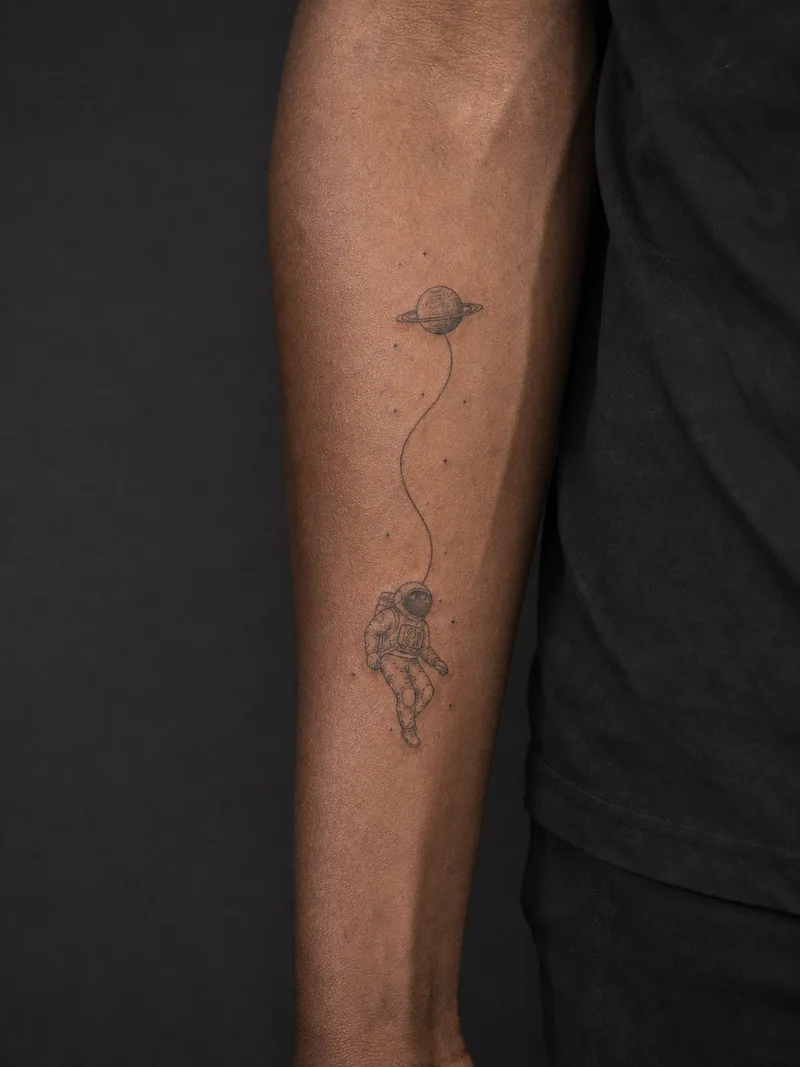 Astronaut tattoo - a floating astronaut connected to a tiny ringed planet by one curved tether