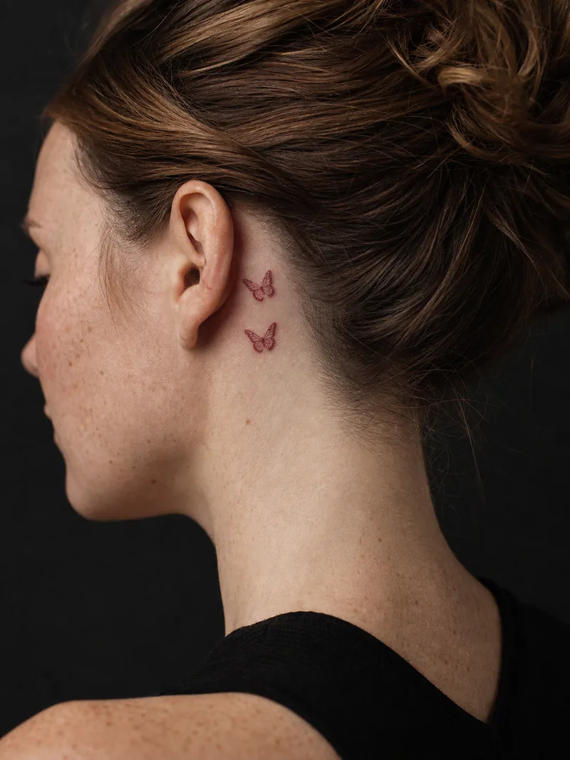 Butterflies behind the ear tattoo - two tiny red butterflies flying upward behind the ear